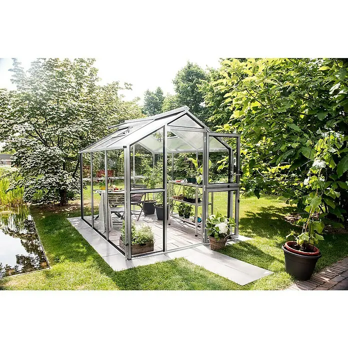 KGT Gewächshaus Galant III - Außenmaß inkl. Dachüberstand (B x T): 216 x 308 cm, Material Seitenwand: Glas, 4 mm, Pressblank - Ein Gewächshaus aus Glas und Metall steht in einem Garten mit Bäumen und einem Teich. Im Inneren des Gewächshauses befinden sich Pflanzen und Möbel.