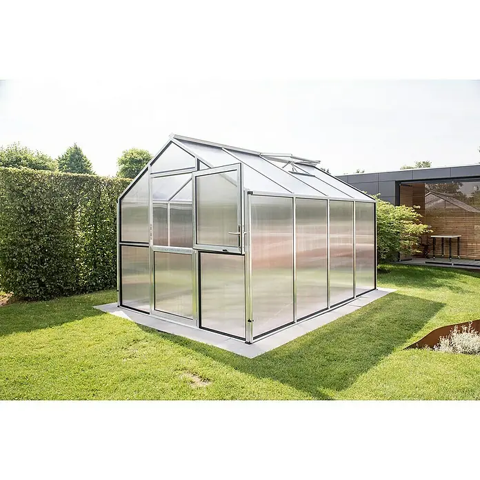 KGT Gewächshaus Novum IV - Außenmaß inkl. Dachüberstand (B x T): 273 x 408 cm, Material Seitenwand: Kunststoff, 10 mm, Pressblank - Nahaufnahme eines Gewächshauses mit Tür auf einem Rasenstück, umgeben von üppigem Grün und einem modernen Gebäude im Hintergrund.