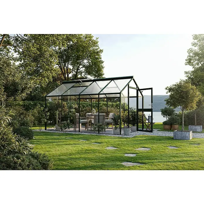 KGT Gewächshaus Novum IV - Außenmaß inkl. Dachüberstand (B x T): 273 x 408 cm, Material Seitenwand: Glas, 4 mm, Tannengrün - Ein grünes Gewächshaus mit einer offenen Tür, umgeben von einem üppigen Garten mit Bäumen und Pflanzen. Im Inneren befindet sich ein Tisch und Stühle.