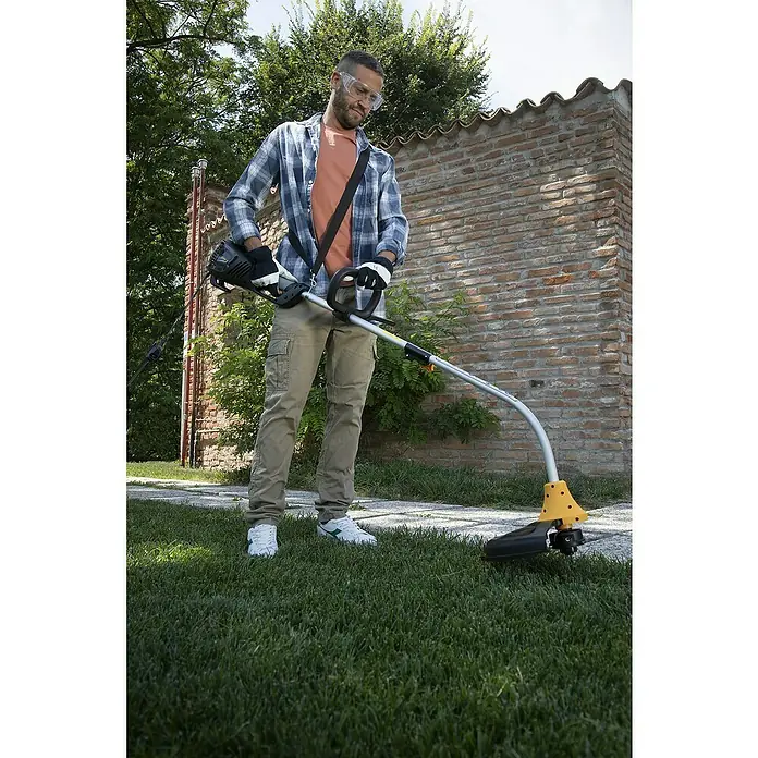 Alpina Recortabordes eléctrico ATR 1.0 EJ - 1.000 W, Ancho de corte hilo: 37 cm - Un hombre usa un cortabordes en el jardín, con gafas de seguridad, guantes y ropa casual para trabajos de jardinería.