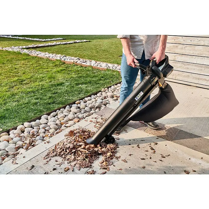 Alpina Soplador de hojas de batería AV20 - 20 V, 2 baterías, 4 Ah, Velocidad de soplado: 55 m/s, Potencia de soplado: 9,6 m³/min - Una persona usa un soplador de hojas negro para quitar hojas en un patio. El soplador de hojas es el foco principal de la imagen.