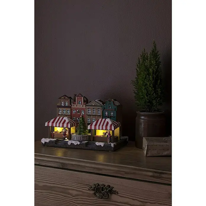 Konstsmide LED-Weihnachtsdorf Stadtszene mit Musik - Bunt, 31,5 x 23 x 23,5 cm, LED, Batteriebetrieben - Ein beleuchtetes Weihnachtsdorf mit Buden und kleinen Figuren steht auf einer Kommode neben einer Topfpflanze.
