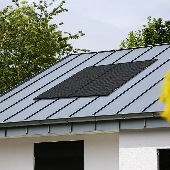 Green Solar Solarmodul-Set 0% MwSt. gem. § 12 Abs. 3 UStG - Nennleistung: 450 W, L x B x H: 176,2 x 113,4 x 3 cm, 1 Stk. - Schwarze Solarmodule auf einem grauen Metalldach eines weißen Hauses, umgeben von grünen Bäumen.