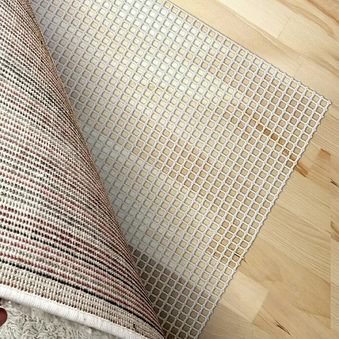 D-c-fix Teppichgleitschutz Loom - 235 x 160 cm - Ein beiger Teppich wird über eine weiße, rutschfeste Unterlage mit Gittermuster auf einem Holzboden ausgerollt.