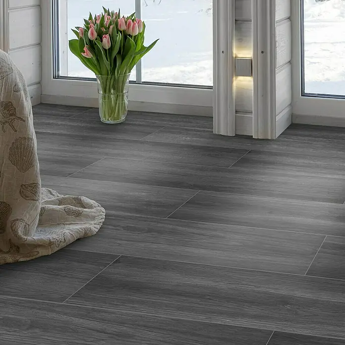 Feinsteinzeugfliese Natura - 22,5 x 90,2 cm, Dark, Matt - Elegante graue Holzbodenfliesen, verlegt in einem Zimmer mit Fenster und Blumenvase. Moderner und stilvoller Look für jeden Raum.