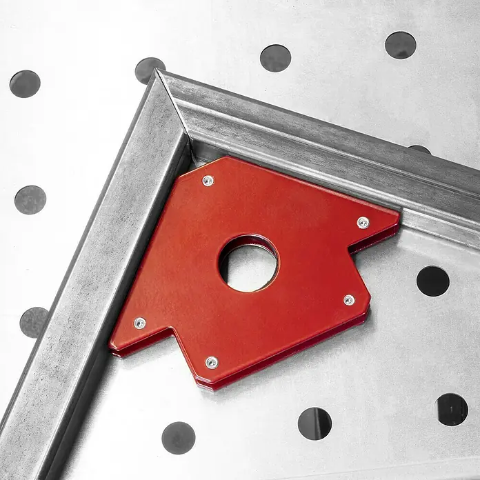 Güde Schweißtisch MST 915 PLUS - L x B: 760 x 500 mm - Nahaufnahme eines roten magnetischen Schweißwinkels, der zwei Metallstücke auf einer perforierten Arbeitsfläche im 90-Grad-Winkel fixiert.
