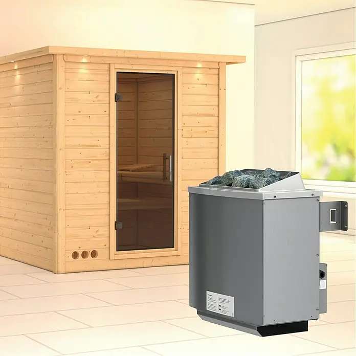 Karibu Elementsauna Mojave - Mit 9 kW Ofen mit integrierter Steuerung, Mit Dachkranz und Beleuchtung, Materialspezifizierung Tür: Ganzglas Graphit, L x B x H: 224 x 210 x 192 cm - Ein modernes Saunaset mit einer hellen Holzsauna und einem grauen Saunaofen mit Steinen. Perfekt für entspannende Wellnessmomente zu Hause.