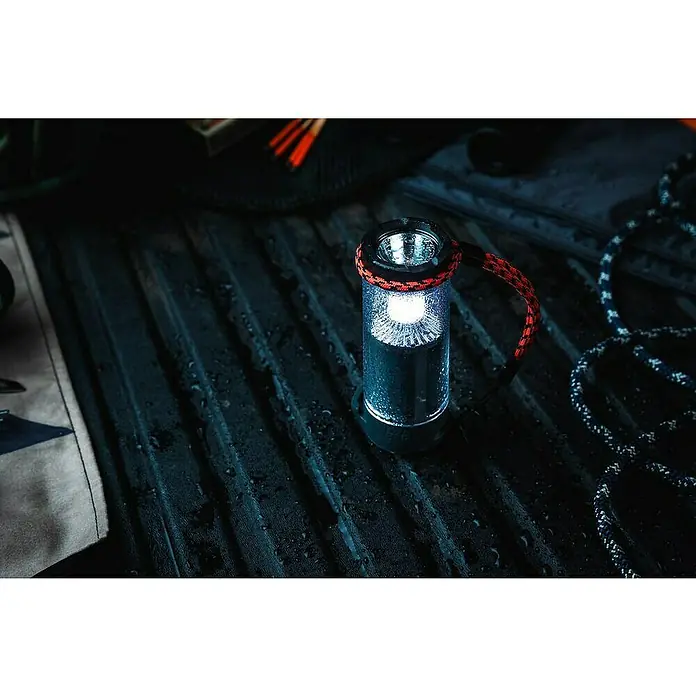 Nebo Tools Lámpara de camping Tempest - IPX7 - Una linterna de exterior brilla intensamente sobre una superficie acanalada, rodeada de objetos útiles para camping o aventuras.