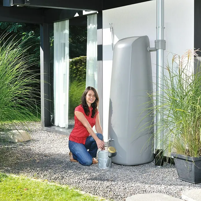 Garantia Classic Line Regenspeicher Elegance - 400 l, Steingrau - Frau befüllt eine Zinkgießkanne an einem grauen Regenspeicher vor einem modernen Haus.