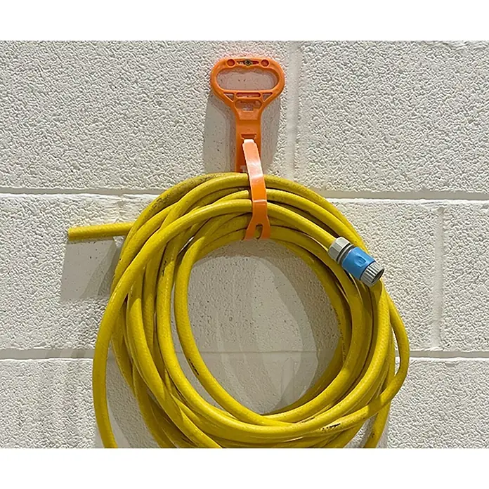 D-Line Kabelverbinder Universalhalterung mit Griff/Aufhängung - Orange - Gelber Gartenschlauch auf einer orangefarbenen Halterung an einer weißen Ziegelwand. Praktische Schlauchaufbewahrung im Außenbereich.