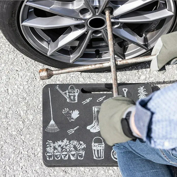 Windhager Motiv Kniekissen - L x B x H: 29 x 39,5 x 2,5 cm - Eine Person wechselt einen Reifen mit einem Radkreuz und kniet auf einem Knieschoner mit Gartenmotiven.