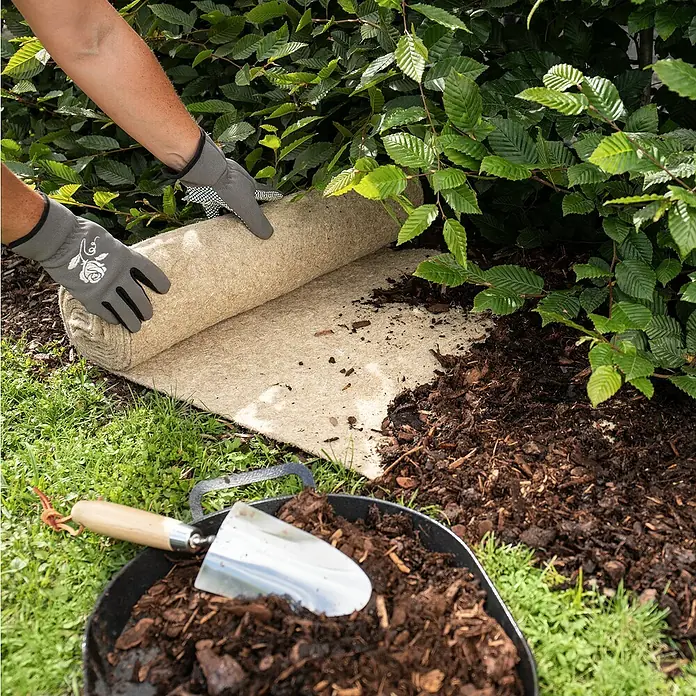 Windhager Unkrautmatte - L x B: 4 x 0,5 m - Ein Gärtner verlegt eine Rolle Unkrautvlies, während Gartengeräte und Mulch bereitstehen, um das Beet vorzubereiten.