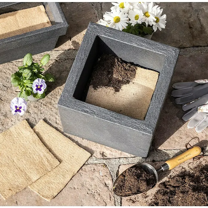 Windhager Jutevliesmatte Jutefilz Wasserspeichermatte - Natur, B x L: 20 x 20 cm - Aufnahme von quadratischen Pflanzgefäßen aus grauem Stein mit Blumenerde, Blumensamen und Gartenwerkzeug auf einer Steinterrasse.