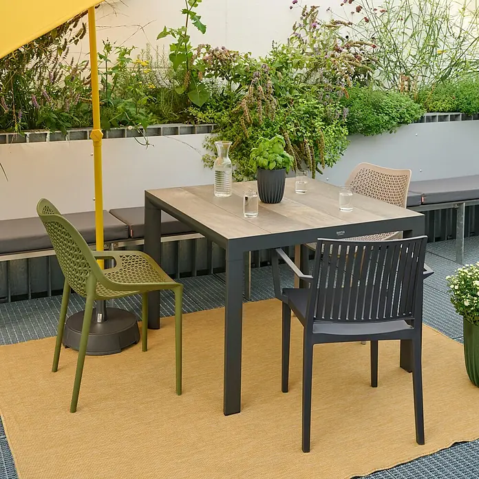 Siena Garden Urban Lights Stapelsessel District - Polypropylen, Stapelbar, Olive, B x T x H: 60 x 50 x 81 cm - Ein Gartentisch mit drei Stühlen in verschiedenen Farben, darunter Grün, Schwarz und Beige, auf einem gelben Teppich im Freien.
