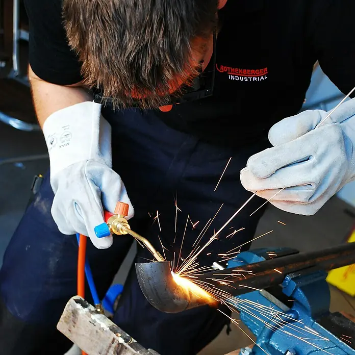 Rothenberger Industrial Equipo de soldadura autógena ROXY KIT Plus - 1.250 °C - Un artesano está soldando una tubería metálica con guantes de protección y equipo de soldadura, con chispas volando.