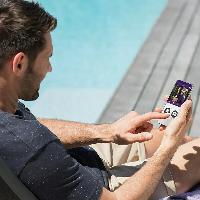 Philips WelcomeEye Türklingel mit Kamera Connect 3 WLAN Komplett-Set - Schwarz - Ein Mann entspannt am Pool und nutzt eine Smart Home App auf seinem Smartphone, um seine Haustür zu überwachen.
