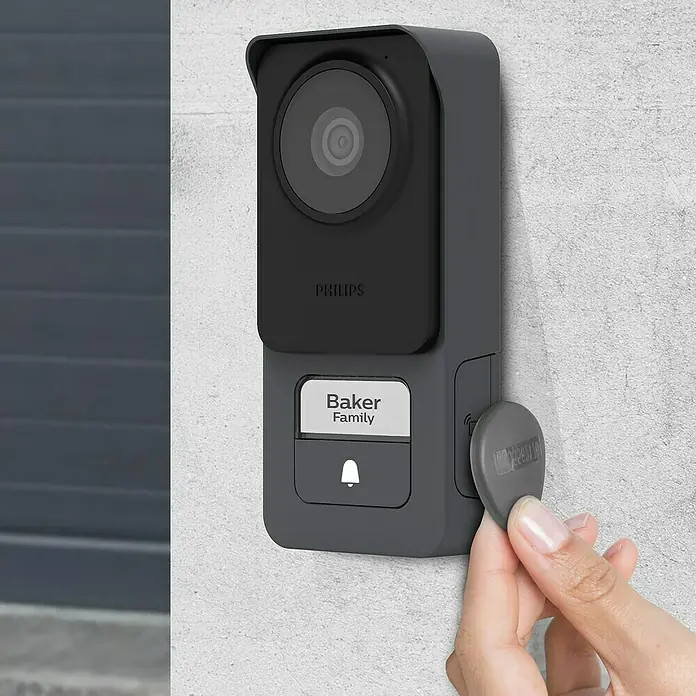 Philips WelcomeEye Türklingel mit Kamera Connect 3 WLAN Komplett-Set - Schwarz - Eine Philips Türklingel mit Kamera, die an einer Wand montiert ist und von einer Hand mit einem Schlüsselanhänger bedient wird.