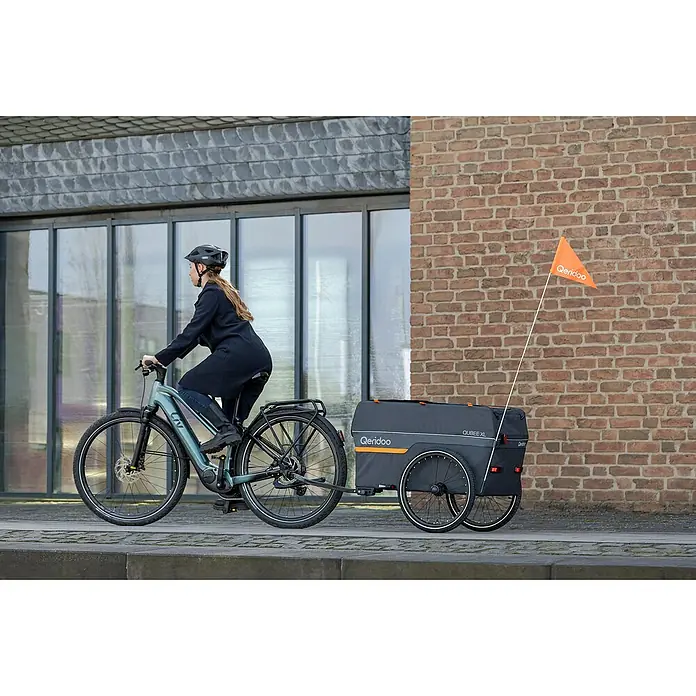 Qeridoo Fahrradanhänger Qubee XL - Max. zulässiges Gesamtgewicht: 60 kg, grau, 220 l - Eine Frau fährt mit einem LIV-Elektrofahrrad, an das ein Qeridoo QUBEEXL-Fahrradanhänger befestigt ist, auf einer Straße.