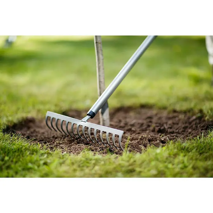 Fiskars Ergonomic Rechen - Aufhängeloch im Griff, Material Gerätekopf: Borstahl - Ein Rechen liegt auf dem Boden neben einem kleinen Baum. Der Rechen wird verwendet, um den Boden um den Baum herum zu glätten.