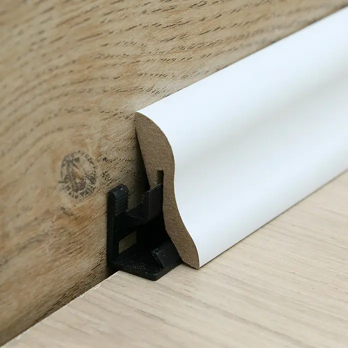 Sockelleiste - 240 x 2,2 x 4 cm, MDF - Nahaufnahme einer weißen Fußleiste mit schwarzer Halterung, die an einer Holzwand und einem Holzboden befestigt ist.