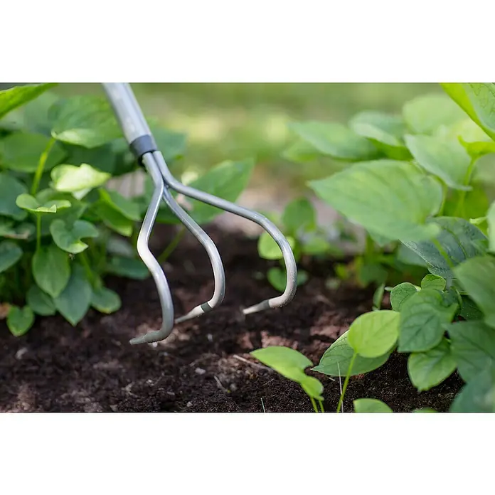 Fiskars Ergonomic Grubber - Material Gerätekopf: Borstahl, Anzahl Zinken: 3, 1 Stk. - Nahaufnahme einer Gartenkralle, die in dunkle Erde zwischen grünen Pflanzen steckt, ideal zum Auflockern und Belüften des Bodens.