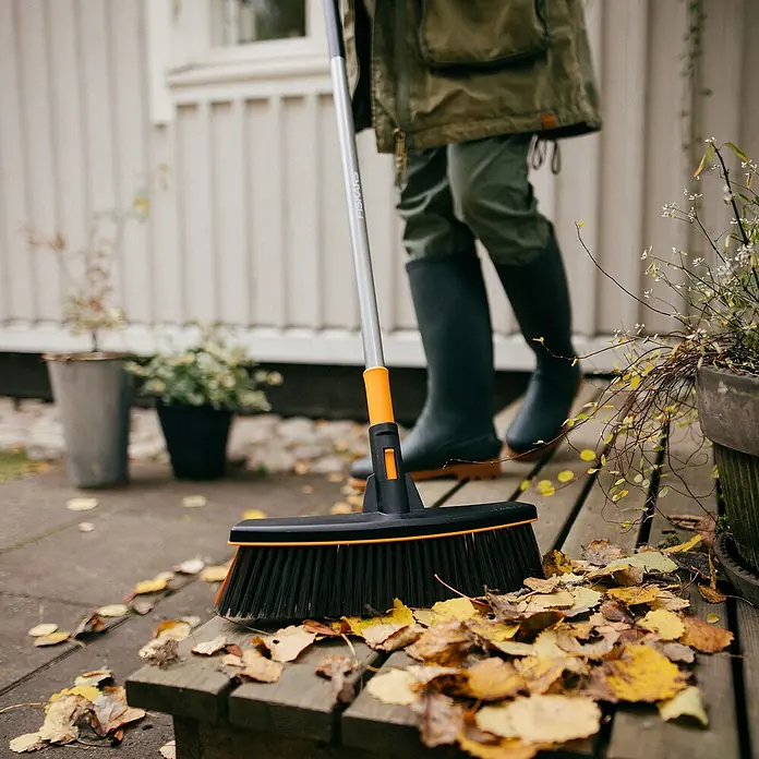 Fiskars Ergonomic Besen - Mit Stiel, Aufhängeloch im Griff - Eine Fiskars Kehrbürste mit Laub und einem Paar Gummistiefel auf einer Terrasse.