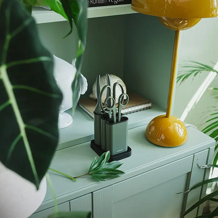 Fiskars Schere - Elegante, freistehende Werkzeughalterung zur praktischen Aufbewahrung, Max. Aststärke: 18 mm - Ein grüner Fiskars Scherenturm mit einer Schere und einem Messer darauf, der auf einer mintfarbenen Kommode neben einer gelben Lampe steht.