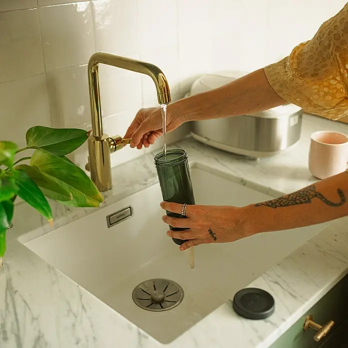 Fiskars Wasserspender - 350 ml, Schwarz - Eine Person befüllt einen dunkelgrünen, selbstbewässernden Pflanzer unter einem goldenen Wasserhahn in einer Spüle mit Marmorarbeitsplatte.