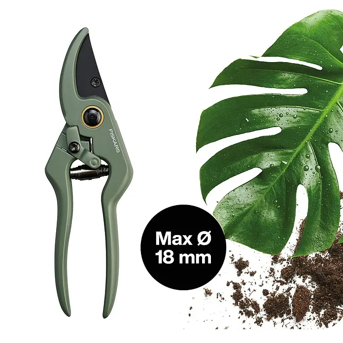 Fiskars Schere - Elegante, freistehende Werkzeughalterung zur praktischen Aufbewahrung, Max. Aststärke: 18 mm - Das Bild zeigt eine grüne Gartenschere der Marke Fiskars neben einem grünen Blatt und Erde. Die Schere hat eine maximale Schnittkapazität von 18 mm.
