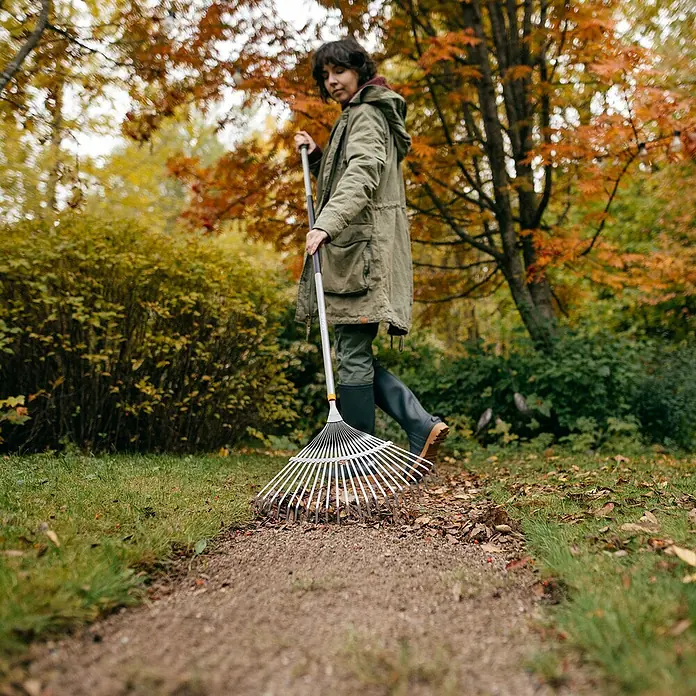Fiskars Ergonomic Fächerbesen - Material Gerätekopf: Borstahl, 1 Stk. - Eine Frau im Park räumt Laub mit einem silbernen Fächerbesen auf einem Weg. Herbstliche Atmosphäre mit bunten Blättern.