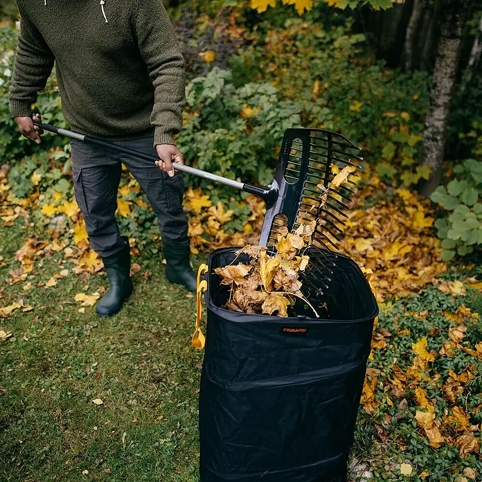 Fiskars Ergonomic Laubschaufel - Material Gerätekopf: Kunststoff, 1 Stk. - Eine Person benutzt einen Fiskars Laubgreifer, um Blätter in einen schwarzen Laubsack zu werfen, umgeben von herbstlichem Laub.