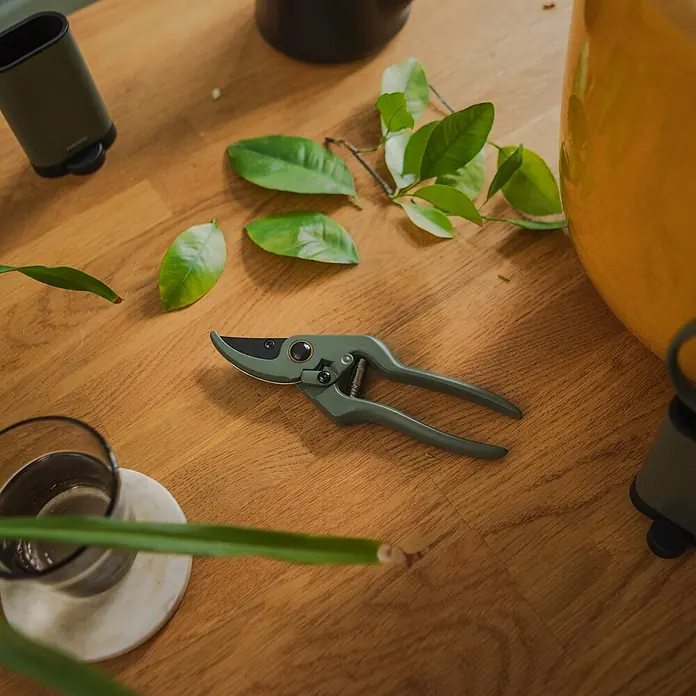 Fiskars Schere - Elegante, freistehende Werkzeughalterung zur praktischen Aufbewahrung, Max. Aststärke: 18 mm - Nahaufnahme einer grünen Gartenschere auf einem Holztisch mit Blättern und Gartenzubehör.