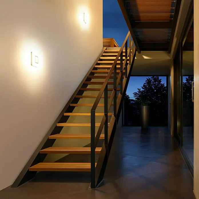 Steinel Sensor-Wand- & Deckenleuchte RS LED M1 S Edelstahl - 8,8 W, L x B x H: 20 x 20 x 5,3 cm, Weiß - Moderne Treppe mit Holzstufen und minimalistischem Design, beleuchtet durch stilvolle Wandleuchten, die ein warmes Ambiente schaffen.