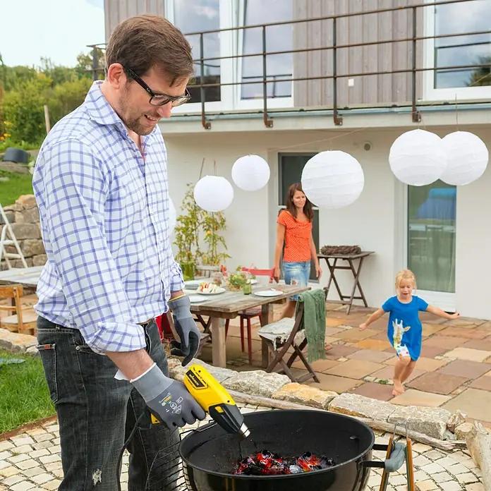 Wagner Heißluftgebläse Furno 300 - Leistungsaufnahme: 1.600 W, Temperaturbereich: 400 °C - 575 °C - Ein Mann zündet mit einem gelben Heißluftfön Kohle in einem Grill an, während seine Familie im Hintergrund zusieht.
