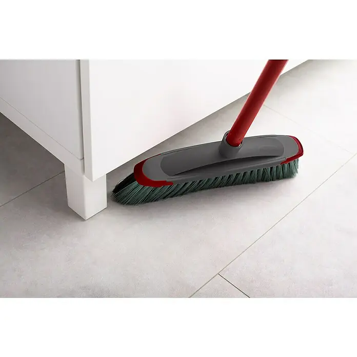 Vileda Zimmerbesen Classica 2in1 Besen - Material Borsten: Recyceltes PET, Breite Besen: 34 cm - Nahaufnahme eines grauen Kehrbesens mit rotem Griff, der unter einem weißen Schrank auf einem hellen Boden kehrt.