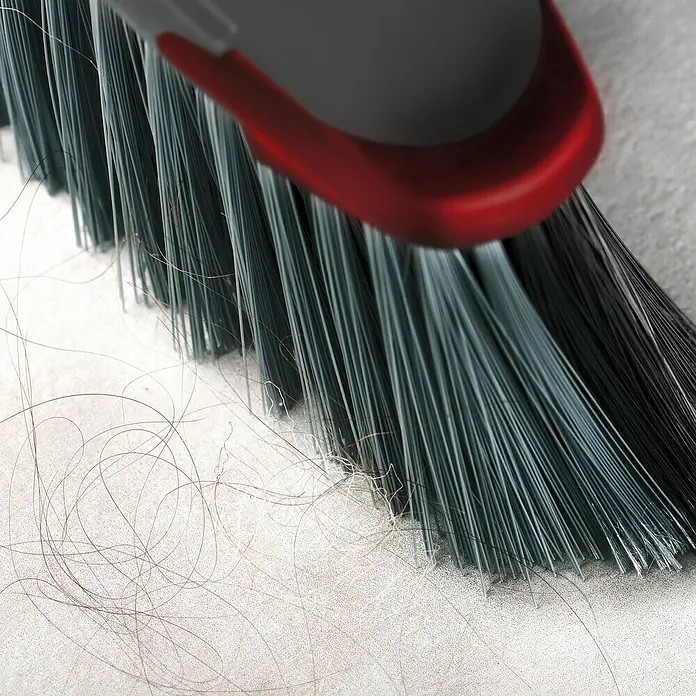 Vileda Zimmerbesen Classica 2in1 Besen - Material Borsten: Recyceltes PET, Breite Besen: 34 cm - Nahaufnahme eines Kehrbesens mit grauen Borsten und roten Akzenten, der Haare auf einer grauen Oberfläche aufkehrt.