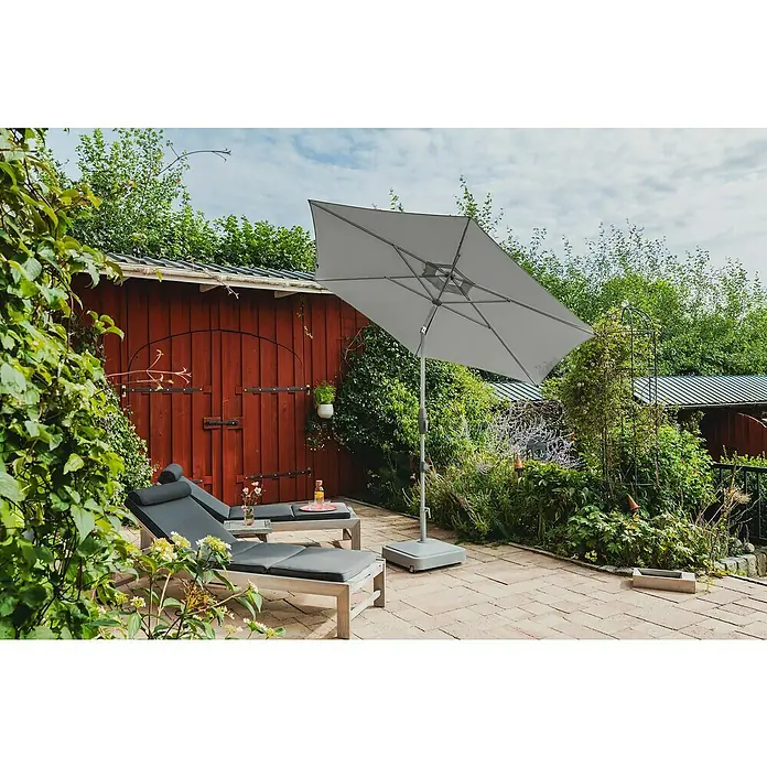 Suncomfort Marktschirm Shell-Turn - Steingrau, Durchmesser Bespannung: 300 cm - Ein grauer, rechteckiger Sonnenschirm beschattet zwei Liegestühle auf einer Terrasse, umgeben von üppigem Grün und einem roten Holzgebäude.