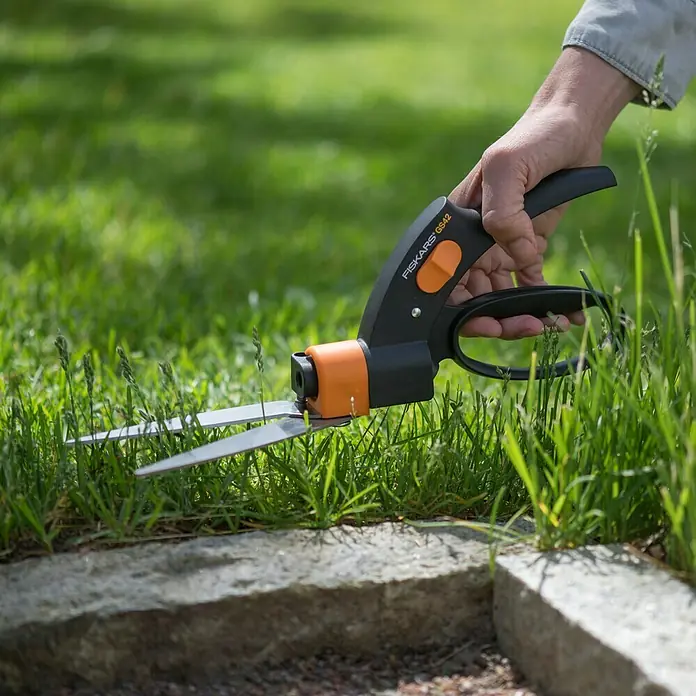 Fiskars Rasenkantenschere Servo System GS42Länge: 32 cm - Bild einer Person, die mit einer Fiskars GS42 Grasschere Gras an einem Steinrand schneidet, Fokus auf die Schere.