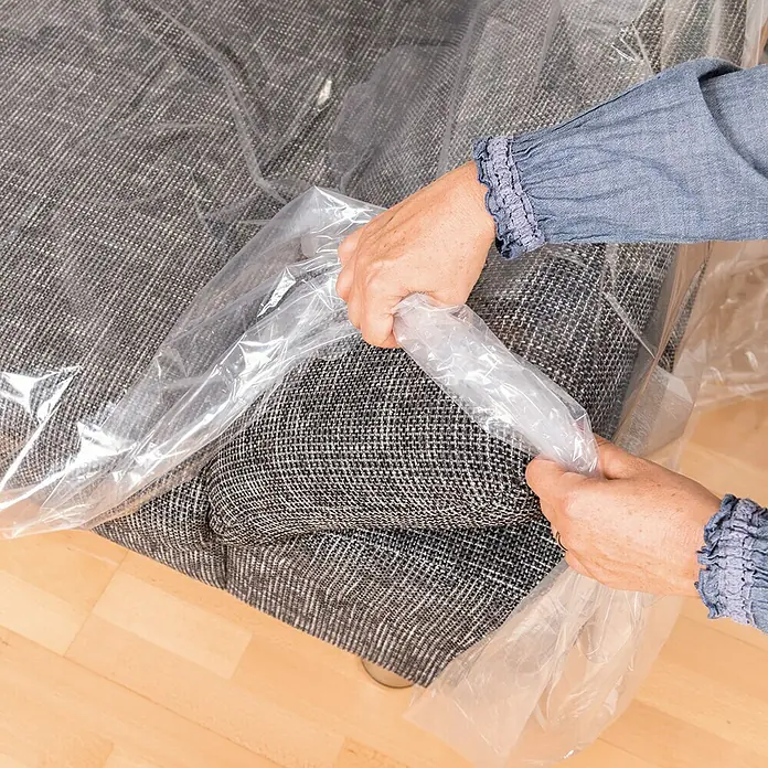 Couchhülle - 300 x 130 cm, Transparent, 2 Stk. - Eine Person verpackt ein graues Sofa sorgfältig in eine transparente Schutzfolie, um es vor Staub und Beschädigungen zu schützen.
