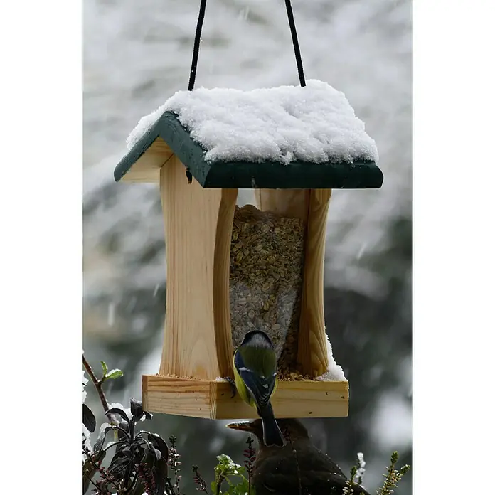 Dobar Futterspender - 16 x 16 x 24 cm, Kiefer, Ausstattung: Mit Silo - Ein mit Schnee bedecktes Vogelhaus mit einem kleinen Vogel, der darauf sitzt und darunter ein weiterer Vogel wartet.