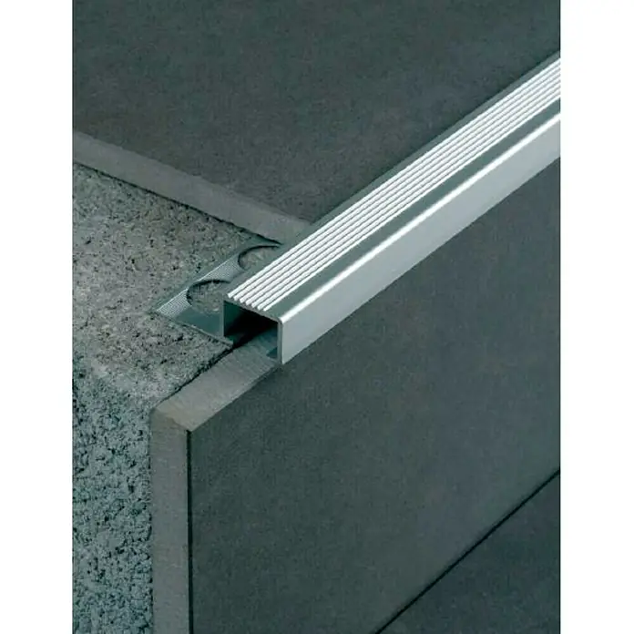Perfil cuadrado para peldaños - Aluminio, Plateado, 13 mm - Una imagen detallada de un borde de escalera metálico de color plateado, incrustado entre dos superficies con texturas contrastantes.
