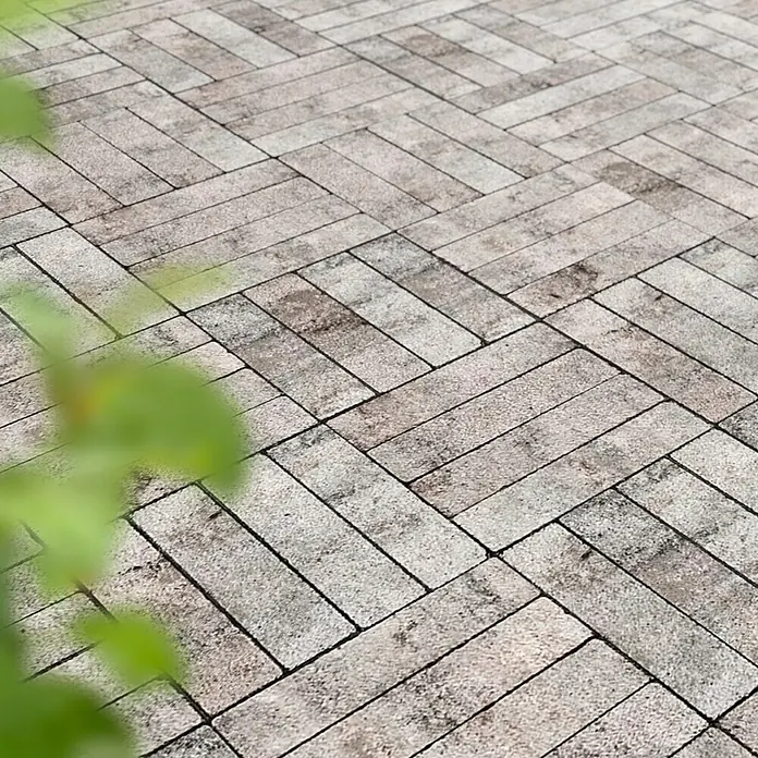 Diephaus Pflasterstein Lorain - Greige, 24 x 6 x 7 cm, Rechteckig - Herringbone-Pflastersteine im Außenbereich, verlegt im eleganten Winkelmuster, mit subtilen Farbvariationen und einem Hauch von Grün im Vordergrund.