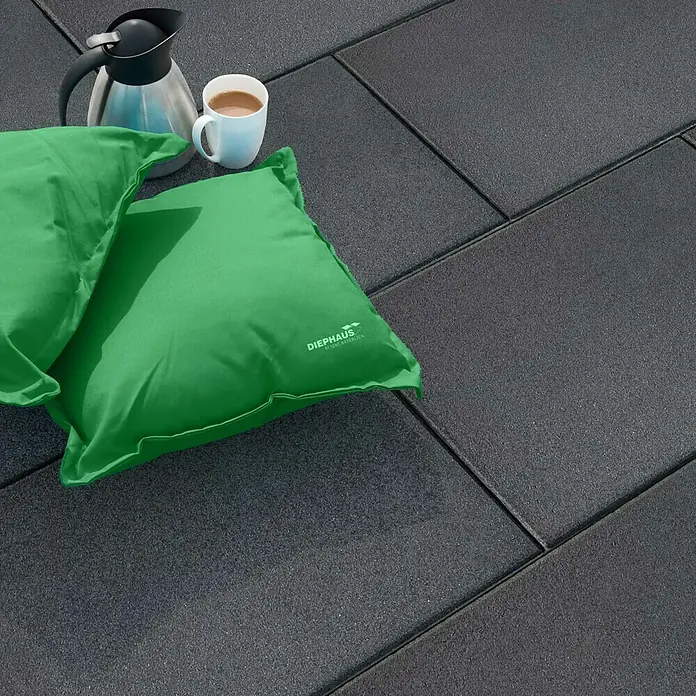 Diephaus Terrassenplatte Norfolg - 60 x 40 x 4 cm, Schwarz/Basalt, Beton - Ein Bild von zwei grünen Kissen, einer Thermoskanne und einer Tasse auf einem grauen Steinboden. Ideal für eine entspannte Pause im Freien.