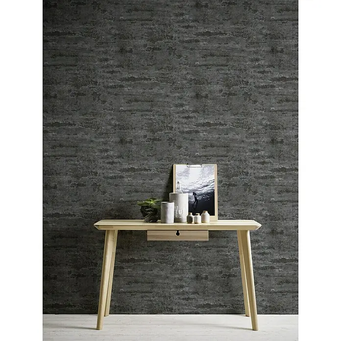 A.S. Création Neue Bude 2.0 ED II Vliestapete Used Ziegel - Schwarz, Steinoptik, 10,05 x 0,53 m - Schreibtisch aus hellem Holz mit Schublade, dekoriert mit Pflanze, Kerzen und einem Bild, vor einer grauen Wand für Büro oder Arbeitszimmer.