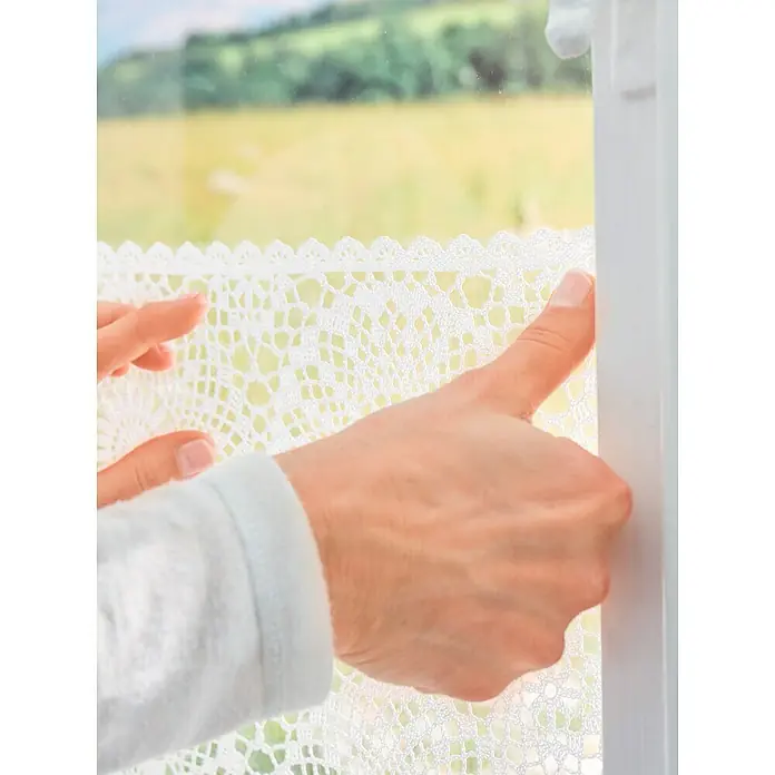 Tesa Powerstrips Cinta engomada - Transparente - Persona instalando una cortina de ganchillo blanca y delicada en la ventana, que proporciona una sutil privacidad y filtrando la luz natural.