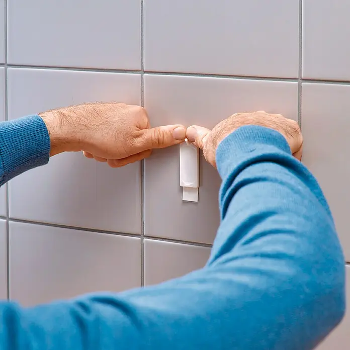 Tesa Clavo adhesivo - Blanco, Carga soportada: 3 kg, 2 ud. - Una persona que fija un gancho autoadhesivo blanco a una pared con azulejos gris claro.