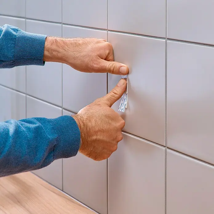 Tesa Tornillo adhesivo ajustable - Rectangular, Carga soportada: 3 kg, 2 ud. - Manos que fijan una tira adhesiva transparente a una pared de azulejos de color gris claro.