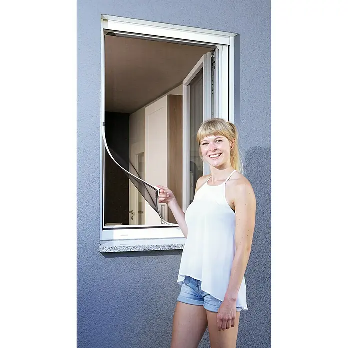 Schellenberg Insect Stop Mosquitera para ventana Magnetic - An x Al: 120 x 120 cm, Montaje de la mosquitera: Fijación magnética, Color bastidor: Blanco - Una mujer abre una ventana con una mosquitera. La mosquitera tiene un borde blanco y protege contra los insectos en la casa.