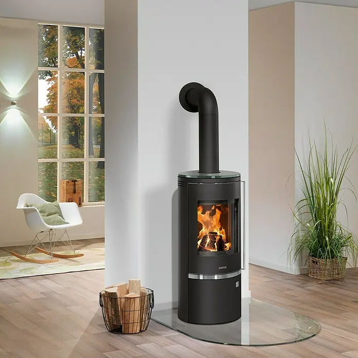 Justus Kaminofen Faro Top 2.0 - 6 kW, Raumheizvermögen: 115 m³, Abdeckung: Glas, Schwarz - Ein moderner Justus Kaminofen mit Glasplatte steht in einem hellen Wohnzimmer und sorgt für eine gemütliche Atmosphäre mit loderndem Feuer.