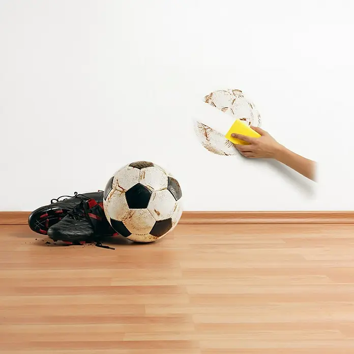 SCHÖNER WOHNEN-Farbe Wandfarbe Sauberweiß - Weiß, 5 l, Matt - Fußballschuhe und -ball liegen auf dem Boden, während eine Hand mit einem Schwamm einen Fleck in Fußballform von der Wand wischt.